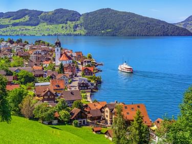 Dorf Beckenried mit Kirchturm am Ufer des Vierwaldstättersees. Raddampfer auf blauem Wasser, grüne Hügel und Berge im Hintergrund.