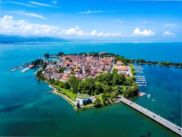 Luftaufnahme der Insel Lindau in Bayern mit historischen Gebäuden, Kirchturm und Yachthäfen, durch eine Brücke mit dem Festland am Bodensee verbunden.