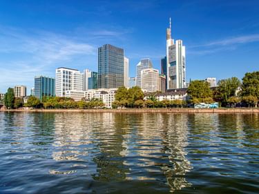 Frankfurt skyline with modern skyscrapers reflected in the Main River. Green trees line the riverbank under a clear blue sky.