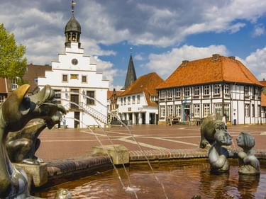 Marktplatz in Lingen mit weißem Rathaus, Brunnen mit Skulpturen und traditionellen Fachwerkhäusern unter bewölktem Himmel.