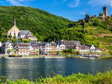 Beilstein an der Mosel mit bunten Häusern, einer Kirche und der Burgruine Metternich auf dem Hügel, umgeben von Weinbergen.
