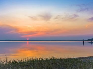 Sonnenuntergang über dem Plauer See mit orange-violettem Himmel, der sich im ruhigen Wasser spiegelt. Gras im Vordergrund, ferne Uferlinie sichtbar.