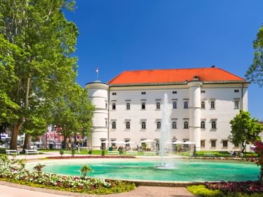 Weißes Renaissance-Schloss Porcia mit orangem Dach und türkisfarbenem Brunnen im Vordergrund, umgeben von bunten Blumenbeeten und Bäumen.