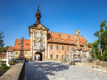 Historisches Bamberger Rathaus mit verzierten Fresken und orangefarbenem Dach auf einer Steinbrücke, flankiert von Barockstatuen unter blauem Himmel.