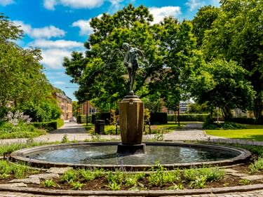 Bronzestatue auf Steinsockel in rundem Brunnen, umgeben von grünem Park mit Bäumen, Wegen und Wohngebäuden in Ängelholm.