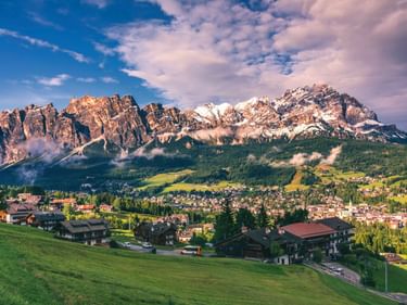 Panoramablick auf Cortina d'Ampezzo in einem Tal mit dramatischen Dolomitengipfeln dahinter, grüne Wiesen im Vordergrund.