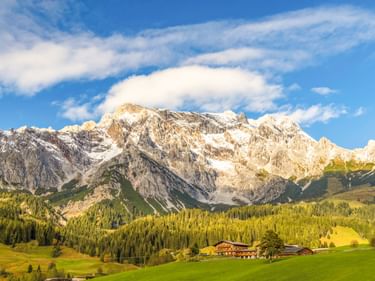 Traditionelle Almhütten auf grünen Wiesen mit dichtem Wald und schneebedeckten Berggipfeln unter blauem Himmel bei Bischofshofen, Österreich.