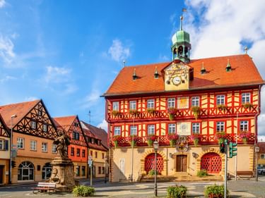 Bad Staffelstein Town Hall with red and yellow timber framing, green tower, and clock. Historic half-timbered buildings and fountain in town square.