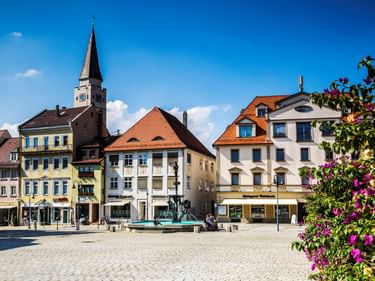 Kopfsteinpflaster-Marktplatz in Ehingen mit bunten historischen Gebäuden, Kirchturm, Brunnen und rosa Blumen im Vordergrund unter blauem Himmel.