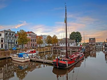 Traditionelle Boote im Hafen von Gorinchem mit bunten niederländischen Häusern am Ufer und einem hochmastigen Segelschiff unter Abendhimmel.