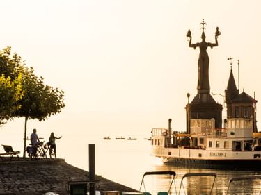 Konstanzer Hafen bei Sonnenuntergang mit der rotierenden Imperia-Statue und historischem Raddampfer. Zwei Personen sitzen an der Uferpromenade unter einem Baum.