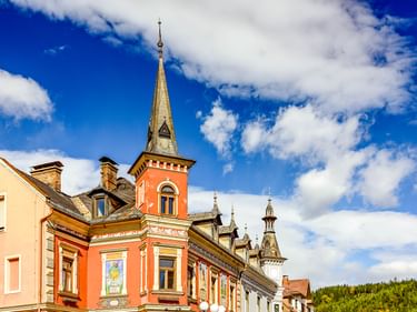 Bunte historische Gebäude in Spittal mit oranger Fassade und hohem Turm vor blauem Himmel. Grüne bewaldete Hügel im Hintergrund.