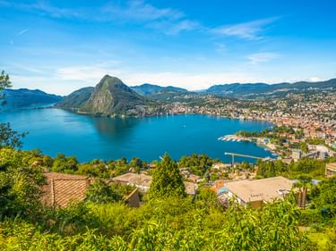 Luftaufnahme von Lugano und dem Luganersee mit dem Berg Monte San Salvatore. Blauer See umgeben von Bergen und Wohngebieten unter klarem Himmel.