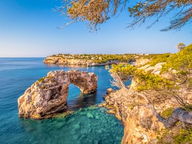 Es Pontàs natürlicher Steinbogen im türkisfarbenen Mittelmeer vor Mallorcas felsiger Küste mit grüner Vegetation und blauem Himmel.