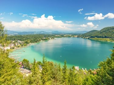 Luftaufnahme des Klopeiner Sees in Österreich mit türkisem Wasser, umgeben von grünen Wäldern und Bergen unter blauem Himmel mit weißen Wolken.