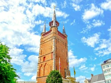 Der historische Belfried von Kortrijk, ein hoher roter Backsteinturm mit mehreren Turmspitzen und goldenen Ornamenten, vor blauem Himmel mit weißen Wolken.