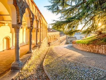 Porticato del Lippomano in Udine mit Steinbögen entlang eines Kopfsteinpflasterwegs. Sonnenlicht fällt durch einen großen Baum rechts.