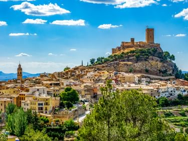 Panoramablick auf Biar, Spanien mit mittelalterlicher Burg auf felsigem Hügel über weißen Häusern. Kirchturm links, Berge im Hintergrund.