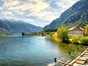 Holzsteg am Hallstätter See in Bad Goisern mit traditionellem Haus am Ufer, umgeben von bewaldeten Bergen unter bewölktem Himmel.