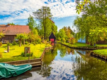 Ruhiger Kanal in Lehde, Spreewald, gesäumt von grünen Wiesen und Bäumen. Traditionelle Häuser und eine kleine Holzkonstruktion säumen den Wasserweg.