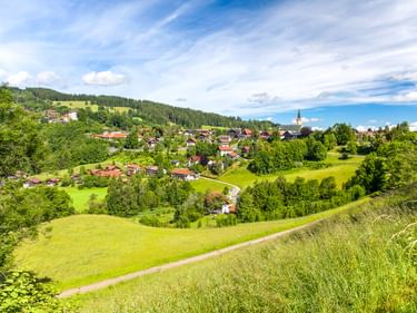 Malerischer Blick auf das Dorf Oberstaufen in grünen Hügeln mit Kirchturm, umgeben von Wäldern und Wiesen unter blauem Himmel.
