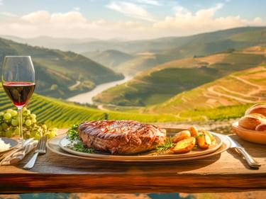 Gegrilltes Steak mit Kartoffeln und Gemüse auf einem Holztisch, mit Rotwein und Brot, mit Blick auf die terrassierten Weinberge des Douro-Tals.