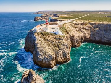 Luftaufnahme des Leuchtturms von Cabo de São Vicente mit rot-weißem Turm auf dramatischen Felsenklippen umgeben von türkisfarbenem Atlantik.