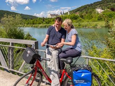 Two cyclists on a bridge near Riedenburg studying a map, with red bicycles and blue panniers. River and green hills with castle visible in background.