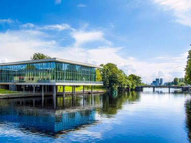 Modernes Glasbibliotheksgebäude auf Stelzen über einem ruhigen Fluss in Halmstad, mit blauem Himmel und grünen Bäumen im Wasser gespiegelt.