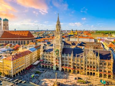 Luftaufnahme des Marienplatzes in München mit dem gotischen Rathausturm, den Kuppeln der Frauenkirche und bunten historischen Gebäuden.