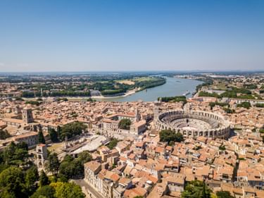 Luftaufnahme von Arles, Frankreich, mit dem antiken römischen Amphitheater, umgeben von Gebäuden mit Terrakottadächern und der Rhône im Hintergrund.