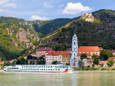 Flusskreuzfahrtschiff auf der Donau vor Dürnstein mit blauem barockem Kirchturm. Historische Gebäude am Ufer, Burgruine auf dem Hügel.