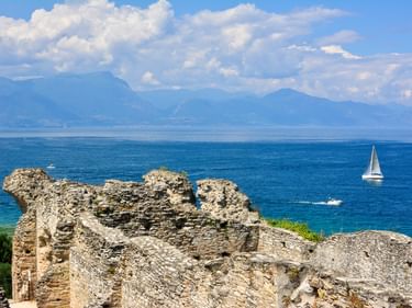 Antike römische Ruinen der Grotte di Catullo mit Steinmauern mit Blick auf den Gardasee. Ein Segelboot gleitet auf blauem Wasser mit Bergen.