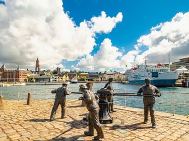 Bronzeskulpturen von Hafenarbeitern im Hafen von Helsingborg mit weißer Fähre und historischen Gebäuden unter blauem Himmel mit weißen Wolken.