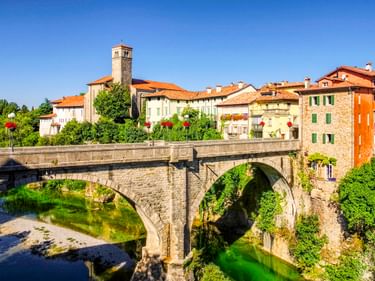 Historische Steinbrücke Ponte del Diavolo über einem grünen Fluss in Cividale del Friuli mit bunten Häusern und Kirchturm.