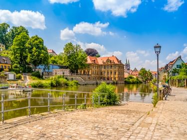 Villa Concordia, ein gelbes Barockgebäude, spiegelt sich in der Regnitz in Bamberg. Kopfsteinpflaster-Promenade mit Geländer und Laterne im Vordergrund.