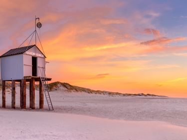 Erhöhter weißer Rettungsturm auf Holzstelzen an einem Sandstrand in Friesland bei Sonnenuntergang. Farbenfroher orange-rosa Himmel mit Dünen im Hintergrund.
