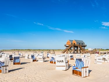 Sandiger Nordseestrand mit nummerierten Strandkörben und einem hölzernen Pfahlbau im Hintergrund unter blauem Himmel.