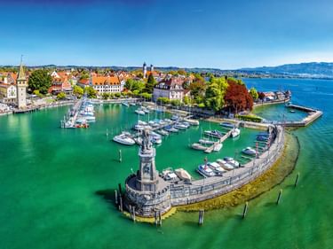 Luftaufnahme des Lindauer Hafens am Bodensee mit ikonischer Löwenstatue auf der Mole, Segelbooten im türkisfarbenen Wasser und historischen Gebäuden.