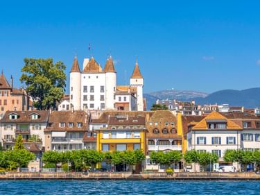 Weißes Schloss Nyon mit Türmen erhebt sich über bunte Ufergebäude am Genfersee. Blauer Himmel und Berge im Hintergrund sichtbar.