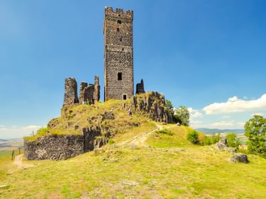 Mittelalterliche Ruine der Burg Hazmburk mit hohem Steinturm auf grasbewachsenem Hügel unter blauem Himmel. Hügellandschaft im Hintergrund.