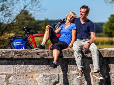 Paar sitzt auf Steinmauer mit rotem Fahrrad während Pause im Altmühltal. Blaue Fahrradtaschen am Rad, ländliche Landschaft im Hintergrund.