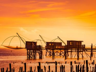 Traditionelle hölzerne Fischerhütten auf Stelzen mit großen Fischernetzen bei Sonnenaufgang in Saint-Nazaire. Orangefarbener Himmel spiegelt sich im ruhigen Wasser.