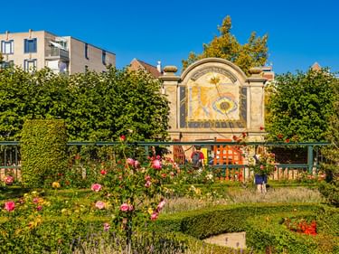 Prinsentuin-Garten in Groningen mit verziertem Sonnenuhr-Monument, bunten Rosenbeeten, geschnittenen Hecken und Besuchern unter blauem Himmel.