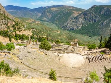 Antikes Steintheater von Delphi mit halbrunden Sitzreihen vor grünen Bergen. Zypressen und Ruinen in der Landschaft sichtbar.
