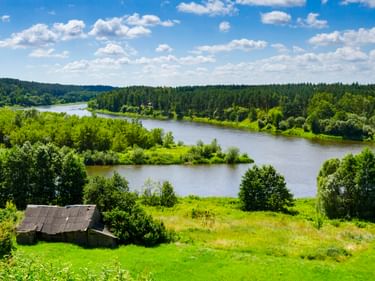Scenic view from Merkine showing a winding river through green forests and meadows. An old wooden barn stands in the foreground under blue sky.