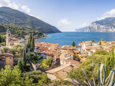 Panoramablick auf Torbole am Gardasee mit Terrakottadächern, Kirchturm, Zypressen und Bergen, die aus dem blauen Seewasser aufragen.