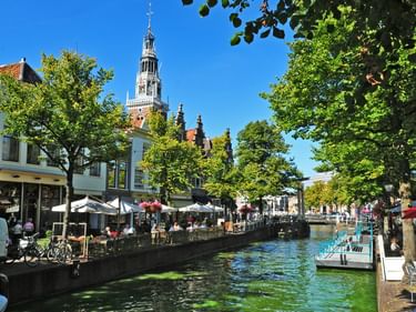 Gracht in Utrecht mit Straßencafés am Wasser, grünen Bäumen und einem hohen Kirchturm im Hintergrund unter blauem Himmel.