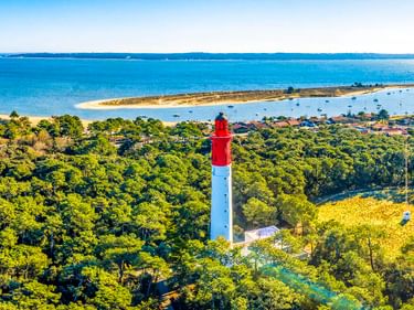 Rot-weißer Leuchtturm von Cap Ferret über dichtem grünen Wald mit Blick auf die Bucht von Arcachon und die Sandküste im Hintergrund.