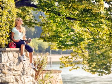 Frau mit Fahrradhelm sitzt auf Steinmauer am Bodensee, liest unter grünen Ästen mit Blick aufs Wasser im Hintergrund.